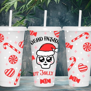 Puede incluir: Tres vasos de plástico blancos con tapas y pajitas. Cada vaso presenta diseños de bastones de caramelo rojos, caramelos en espiral y corazones. Un vaso tiene una calavera con un gorro de Papá Noel con el texto "DEAD INSIDE BUT JOLLY."