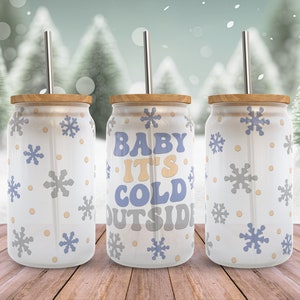 May include: Three white stainless steel tumblers with a wood lid and a straw. The tumblers have a snowflake pattern and the middle tumbler says "Baby It's Cold Outside".