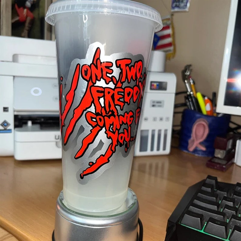 K&ouml;nnte beinhalten: Ein durchsichtiger Plastikbecher mit einer silbernen und roten Grafik, auf der "One Two Freddy Coming For You..." steht. Der Becher steht auf einem silbernen Sockel.