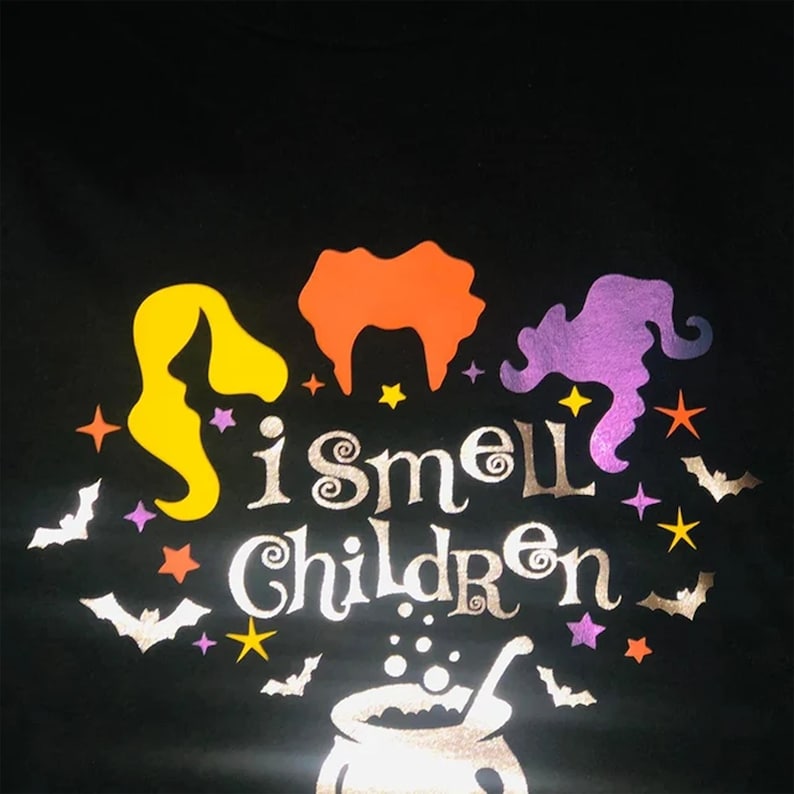 May include: A black t-shirt with a silver glitter design featuring three witches with the text "I smell children" and a cauldron with bubbling liquid.