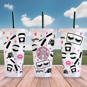 May include: Three white reusable coffee cups with black lids and green straws. Each cup has a different design featuring black and pink makeup-themed illustrations, including eyelashes, lips, mascara, and eyeliner. The text "gorgeous" appears on two of the cups.
