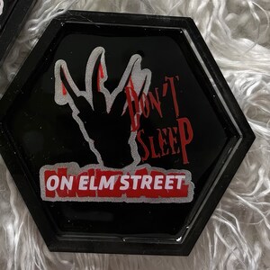 K&ouml;nnte beinhalten: Schwarzer sechseckiger Untersetzer mit einer roten und wei&szlig;en Grafik einer Hand mit vier Fingern, dem Text "Don't Sleep On Elm Street" und einem roten Rand.