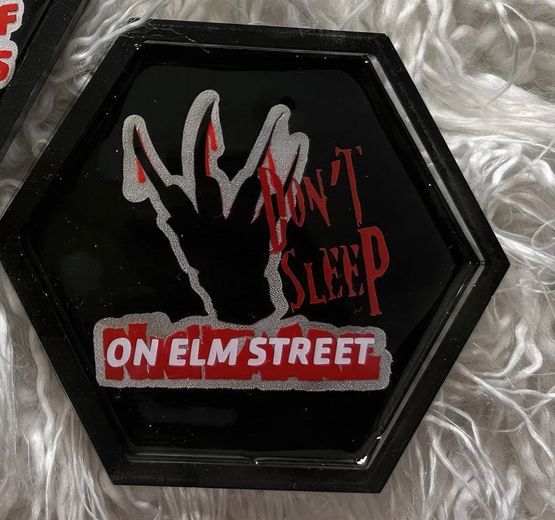 K&ouml;nnte beinhalten: Schwarzer sechseckiger Untersetzer mit einer roten und wei&szlig;en Grafik einer Hand mit vier Fingern, dem Text "Don't Sleep On Elm Street" und einem roten Rand.