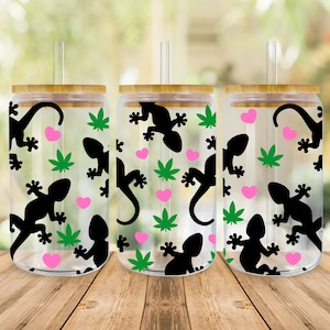 Pode incluir: Três copos de vidro transparentes com tampas de bambu e canudos pretos. Cada copo apresenta um padrão repetido de geckos pretos, folhas de maconha verdes e corações cor-de-rosa.
