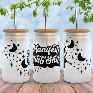 K&ouml;nnte beinhalten: Drei wei&szlig;e Becher mit schwarzen Designs. Der Becher in der Mitte hat den Text "Manifest that Shit" in einer schwarzen, stilisierten Schrift. Die Becher auf beiden Seiten haben ein Halbmond- und Sternenmuster. Jeder Becher hat einen Bambusdeckel und einen Edelstahlstrohhalm.