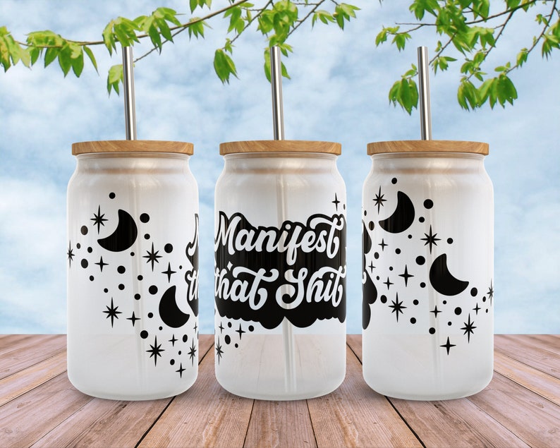 K&ouml;nnte beinhalten: Drei wei&szlig;e Becher mit schwarzen Designs. Der Becher in der Mitte hat den Text "Manifest that Shit" in einer schwarzen, stilisierten Schrift. Die Becher auf beiden Seiten haben ein Halbmond- und Sternenmuster. Jeder Becher hat einen Bambusdeckel und einen Edelstahlstrohhalm.