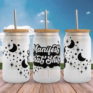 K&ouml;nnte beinhalten: Drei wei&szlig;e Glasbecher mit schwarzen Bambusdeckeln und schwarzen Strohhalmen. Die Becher haben ein schwarz-wei&szlig;es Design mit Monden und Sternen. Der mittlere Becher hat den Text "Manifest that Shit" in Schwarz.