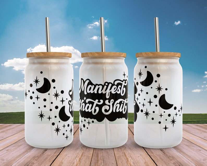 K&ouml;nnte beinhalten: Drei wei&szlig;e Glasbecher mit schwarzen Bambusdeckeln und schwarzen Strohhalmen. Die Becher haben ein schwarz-wei&szlig;es Design mit Monden und Sternen. Der mittlere Becher hat den Text "Manifest that Shit" in Schwarz.