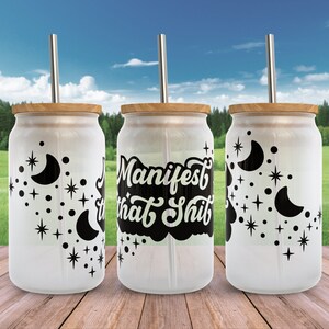 K&ouml;nnte beinhalten: Drei wei&szlig;e wiederverwendbare Becher mit schwarzen Deckeln und Strohhalmen. Die Becher haben ein schwarz-wei&szlig;es Design mit Sternen und Halbmonden. Der mittlere Becher tr&auml;gt den Text "Manifest that Shit" in Schwarz.