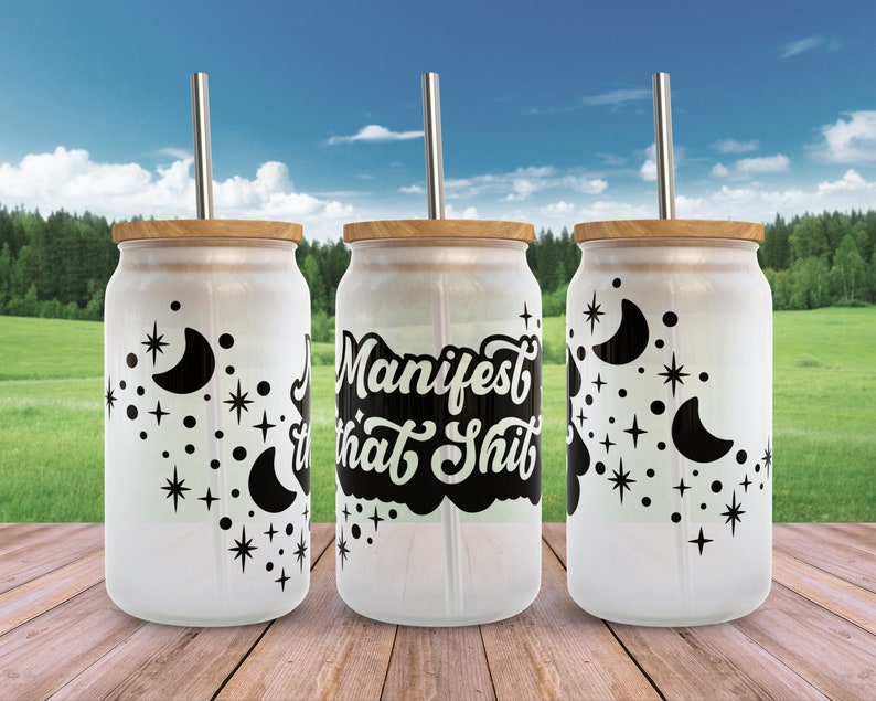K&ouml;nnte beinhalten: Drei wei&szlig;e wiederverwendbare Becher mit schwarzen Deckeln und Strohhalmen. Die Becher haben ein schwarz-wei&szlig;es Design mit Sternen und Halbmonden. Der mittlere Becher tr&auml;gt den Text "Manifest that Shit" in Schwarz.