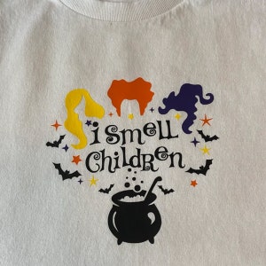 May include: White t-shirt with a black cauldron, three witches' silhouettes, and the text "i smell children" in black, yellow, orange, and purple.