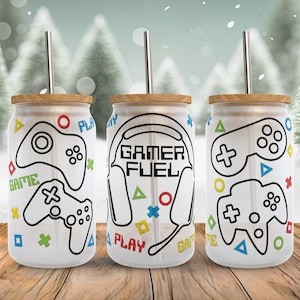 May include: Three white tumblers with lids and straws. Each tumbler has a different design featuring a video game controller, the words "Gamer Fuel", and various geometric shapes in blue, green, yellow, red, and orange.
