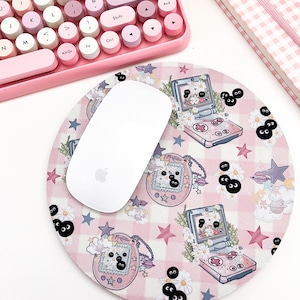 May include: A pink and white checkered mouse pad with a repeating pattern of a pink and white handheld game console, stars, and black eyes. A white wireless mouse is on top of the mouse pad.