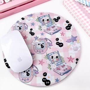 May include: A pink and white checkered mouse pad with a cartoon design featuring a pink handheld game console, stars, and black eyes. The mouse pad is round and has a white mouse on top of it.