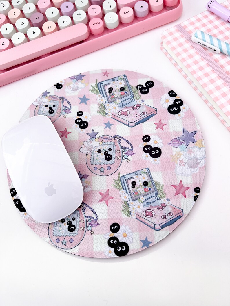 May include: A pink and white checkered mouse pad with a cartoon design featuring a pink handheld game console, stars, and black eyes. The mouse pad is round and has a white mouse on top of it.