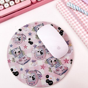 May include: A pink and white round mousepad with a repeating pattern of pink and white checkered squares, stars, and cartoon images of a handheld video game console with a black and white character on the screen. The mousepad has a white mouse on top of it.