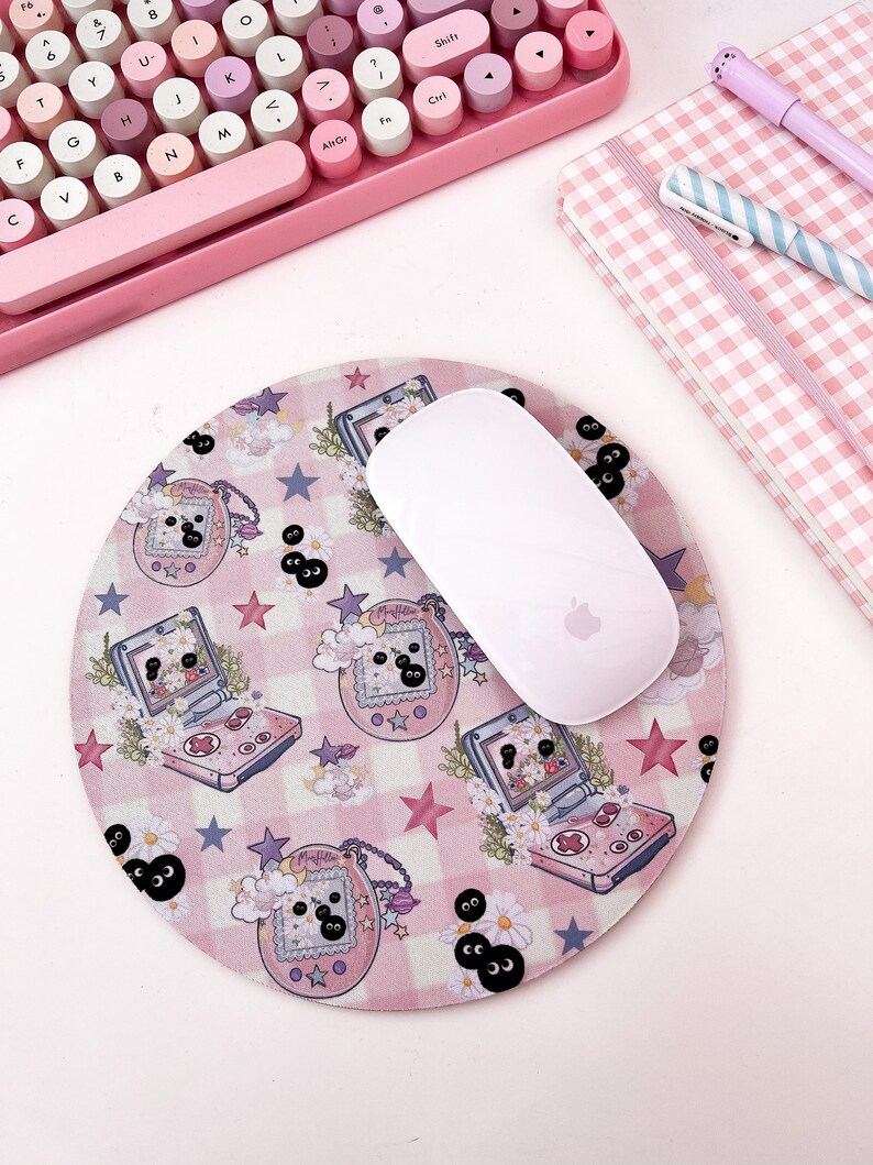 May include: A pink and white round mousepad with a repeating pattern of pink and white checkered squares, stars, and cartoon images of a handheld video game console with a black and white character on the screen. The mousepad has a white mouse on top of it.