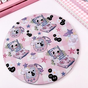 May include: A pink and white round mousepad with a checkered pattern and a repeating design of cartoon handheld video game consoles and stars. The consoles are pink and white with black buttons and screens. The stars are pink and white. The background is a pink and white checkered pattern.