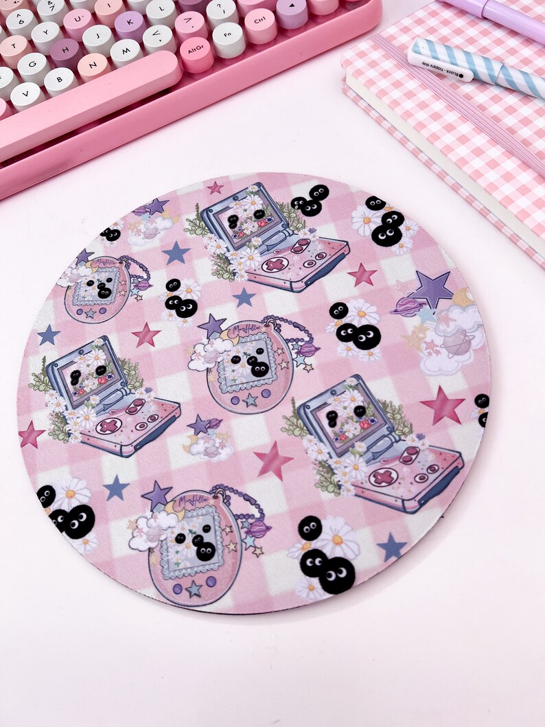 May include: A pink and white round mousepad with a checkered pattern and a repeating design of cartoon handheld video game consoles and stars. The consoles are pink and white with black buttons and screens. The stars are pink and white. The background is a pink and white checkered pattern.