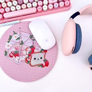 May include: A pink and white mouse pad with a grid pattern. The mouse pad features a cartoon illustration of a glass of milk with strawberries, a slice of bread with strawberries, and a bear face.