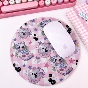May include: A pink and white mouse pad with a checkered pattern and a repeating design of a pink and white handheld video game console with a black and white screen and a black and white character on the screen. The mouse pad also has a repeating design of black and white eyes with white eyelashes. The mouse pad is on a white surface with a pink keyboard and a pink and white checkered notebook in the background.