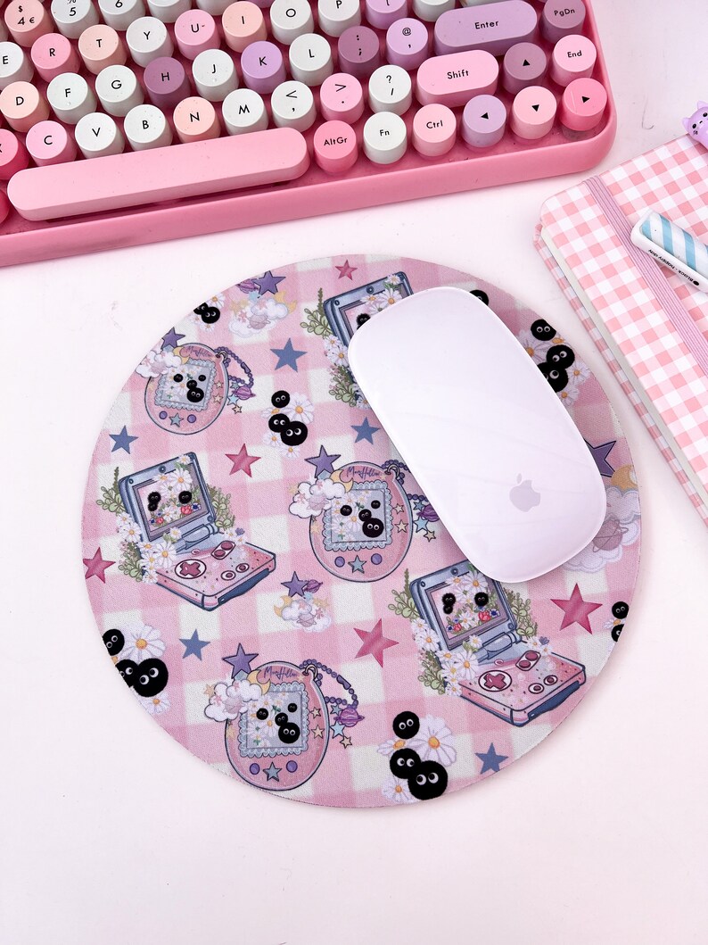 May include: A pink and white mouse pad with a checkered pattern and a repeating design of a pink and white handheld video game console with a black and white screen and a black and white character on the screen. The mouse pad also has a repeating design of black and white eyes with white eyelashes. The mouse pad is on a white surface with a pink keyboard and a pink and white checkered notebook in the background.