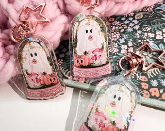 May include: Clear acrylic keychains featuring a whimsical ghost design inside a snow globe. The keychains have a pink base with a lace design, a rose gold star-shaped clasp, and a pink and white ghost illustration with pumpkins and mushrooms.