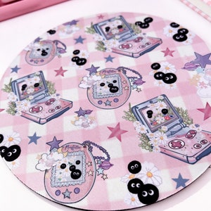May include: A pink and white checkered mousepad with a repeating pattern of a pink and white handheld game console, a pink and white digital pet, and black eyes. The mousepad is round and has a black border.
