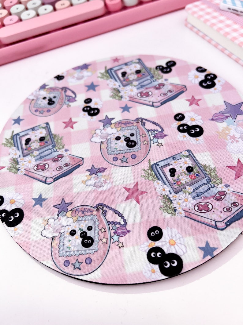 May include: A pink and white checkered mousepad with a repeating pattern of a pink and white handheld game console, a pink and white digital pet, and black eyes. The mousepad is round and has a black border.