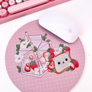 May include: A pink and white mouse pad with a grid pattern. The mouse pad features a cartoon illustration of a bear wearing a strawberry crown, a glass of milk with strawberries, and a straw.