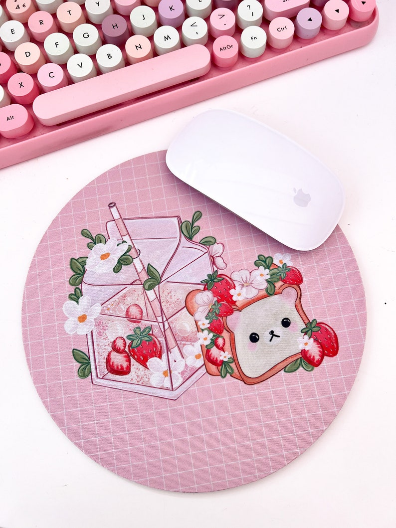 May include: A pink and white mouse pad with a grid pattern. The mouse pad features a cartoon illustration of a bear wearing a strawberry crown, a glass of milk with strawberries, and a straw.
