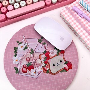 May include: A pink and white mouse pad with a grid pattern. The mouse pad features a cartoon illustration of a glass of milk with a straw, strawberries, and a piece of toast with a bear face.