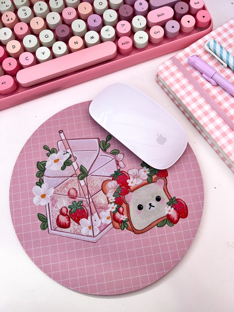 May include: A pink and white mouse pad with a grid pattern. The mouse pad features a cartoon illustration of a glass of milk with a straw, strawberries, and a piece of toast with a bear face.