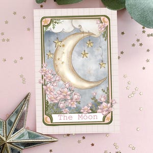 May include: A tarot card featuring a crescent moon and stars against a blue sky. The card is bordered in green with the words "The Moon" printed in pink at the bottom.
