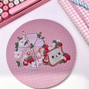 May include: A pink and white round mousepad with a grid pattern. The mousepad features a cartoon illustration of a bear-shaped piece of toast with strawberries and a glass of strawberry milk with a straw.