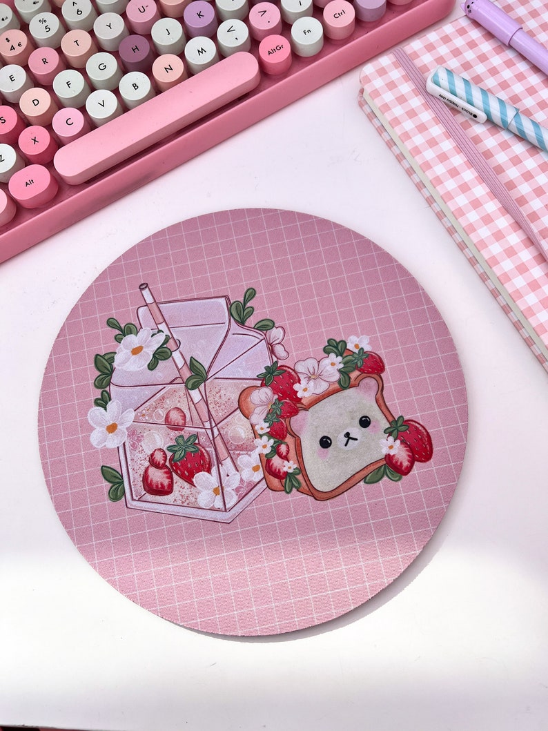 May include: A pink and white round mousepad with a grid pattern. The mousepad features a cartoon illustration of a bear-shaped piece of toast with strawberries and a glass of strawberry milk with a straw.