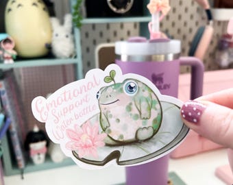 May include: A white sticker featuring a green frog perched on a lily pad. The sticker includes pink flowers and the text "Emotional Support Water Bottle".