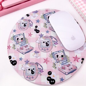 May include: A pink and white mousepad with a checkered pattern and a repeating design of a pink and blue handheld game console, a pink and purple charm with the word "My Melody" on it, and black and white eyes. A white wireless mouse is sitting on the mousepad.