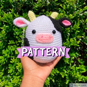 May include: A crocheted cow amigurumi toy with black and white spots, pink ears, and a yellow nose. The toy is being held in a hand with the word "PATTERN" written in pink letters on a banner.