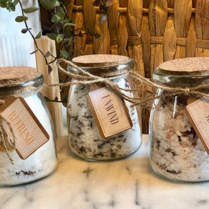 May include: Three glass jars with cork lids filled with bath salts. The jars are tied with twine and have paper tags with the words "Refresh", "Unwind", and "Soak" printed on them.