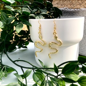 May include: A pair of gold-coloured snake earrings. The earrings have a detailed, textured design and are dangling. They are displayed in front of a white ceramic container, with green leafy plants in the foreground.