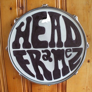 Puede incluir: Un parche de tambor redondo con las palabras "HEAD FRAMEZ" en letras grandes y estilizadas de color marrón oscuro. El parche de tambor está sobre un fondo de madera clara. El tambor tiene un borde de metal plateado.