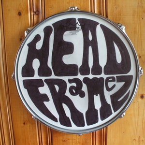 Pode incluir: Uma pele de tambor redonda com as palavras "HEAD FRAMEZ" em letras grandes e pretas estilizadas. A pele do tambor é branca com uma borda de metal prateado. O fundo é uma parede de madeira.