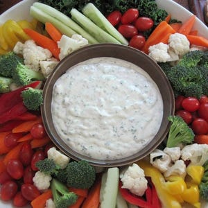 May include: A white platter with a variety of raw vegetables, including carrots, celery, cauliflower, broccoli, bell peppers, and cherry tomatoes. A brown bowl filled with a white creamy dip is in the center of the platter.