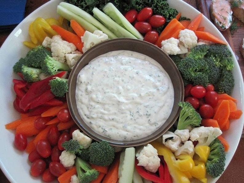 May include: A white platter with a variety of raw vegetables, including carrots, celery, cauliflower, broccoli, bell peppers, and cherry tomatoes. A brown bowl filled with a white creamy dip is in the center of the platter.