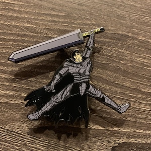 May include: A metal pin featuring a character from the manga series Berserk. The character is depicted in a dynamic pose, holding a large sword overhead. The pin has a black, gray, and gold color scheme.