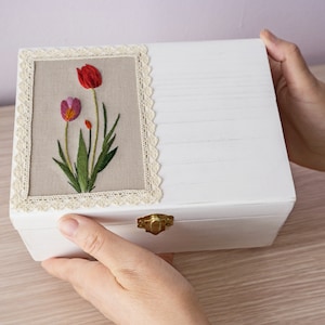 Può includere: Scatola di legno bianca con coperchio incernierato e chiusura dorata. Il coperchio presenta un pannello rettangolare ricamato con un motivo floreale di tre tulipani rosa e rossi su uno sfondo di lino beige. Il pannello è incorniciato da pizzo bianco.