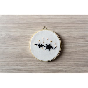 Può includere: Un telaio da ricamo rotondo con sfondo in tessuto bianco, con un disegno di stelle nere. Le stelle sono collegate da un filo nero, con piccole perline dorate. Il telaio ha una cornice in legno e un anello dorato per appenderlo.
