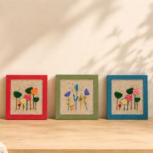 Può includere: Tre opere d'arte floreali incorniciate con cornici rosse, verdi e blu. Ognuna presenta un colorato disegno floreale ricamato su uno sfondo neutro. L'opera d'arte è esposta su una superficie di legno con piante decorative e una candela.