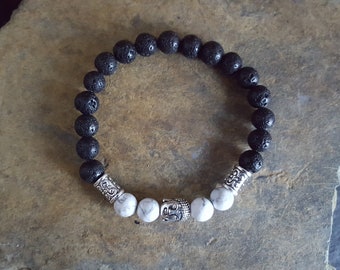 May include: A beaded bracelet featuring a series of round, porous black lava beads and four white, grey-veined marble beads. The design includes a central silver-toned metal Buddha head charm flanked by two ornate silver-toned cylindrical spacers.